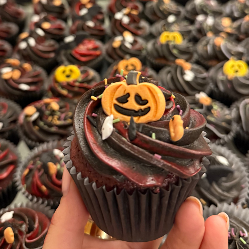 Halloween-themed cupcake with a jack-o'-lantern face held above a large pile of similar cupcakes.